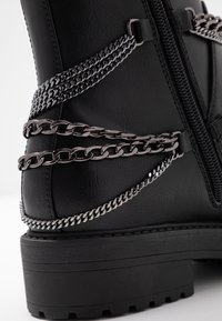 Botín de cuero negro que presenta cadenas plateadas alrededor de la parte superior. Suela gruesa negra con detalles texturizados y cremallera lateral.