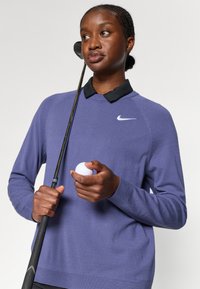Purpuros adītu džemperi ar melnu kreklišu ar apkakli apakšā, ar balto Nike logo. Individuāls tur golfa nūju un golfa bumbu.