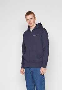 Felpa con zip di colore blu navy, con cappuccio regolabile, tasche frontali e logo GANT 1949 grigio sul petto. Tessuto morbido con una texture liscia.