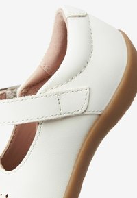 Vit läder sko med rundad tå, sydda detaljer, ett kardborreband och en beige gummisula. Har en utskärningdesign.