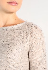 Suéter de punto metálico con cuello redondo, color beige claro y lentejuelas plateadas dispersas, creando un efecto texturizado y brillante.