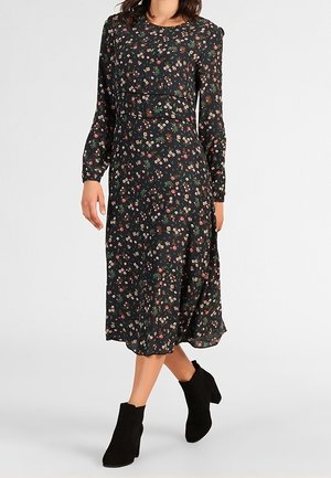 Kvinde iført en sort midi-kjole med lange ærmer og et lille farverigt blomsterprint samt sorte ankelstøvler, stående med det ene ben krydset.