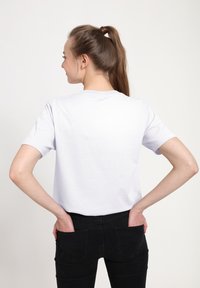 Bílý bavlněný tričko s krátkými rukávy, kulatým výstřihem, jednoduchým designem, nošené s černými džínami, ruce vložené do zadních kapes.