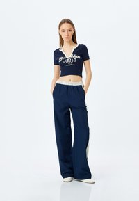 Top corto azul marino con letras blancas y escote, combinado con pantalones de pierna ancha azul marino con cintura elástica y acentos de rayas laterales.