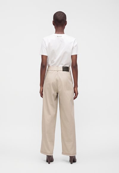 Personne aux cheveux courts portant une chemise blanche à manches courtes et un pantalon beige à jambes larges, debout devant un fond blanc uni.