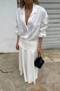 Balti lino marškiniai su laisvais rankovėmis ir dviem priekiniuose kišenėse, derinami su balta sijonu, turinčiu kreivą juosmenį, laikomi juoda rankine.