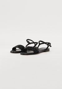 Svart flate sandaler med naglede stropper, minimalistisk design og justerbar ankelrem. Laget hovedsakelig av syntetiske materialer.