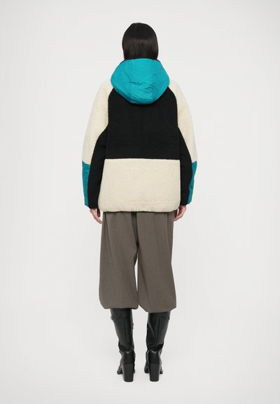 Jachetă pufoasă, cu blocuri de culoare în nuanțe de negru, crem și teal. Are glugă, croială lejeră și material texturat. Este purtată cu pantaloni evazați gri și botine negre.