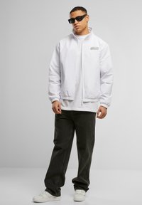 White zip-up jacket with elastic cuffs and a logo, worn over a white t-shirt. Dark gray jeans and white sneakers complete the outfit.