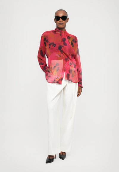 Doorzichtige bloemenblouse in levendig roze met donkere patronen, gecombineerd met witte wijde broek en zwarte puntige hakken.