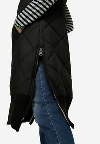 Schwarzige, gesteppte, puffy Weste mit seitlichen Reißverschlüssen, ausgestattet mit einem gerippten Saum. Über einem gestreiften Langarmoberteil getragen und mit blauen Jeans kombiniert.
