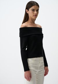 Zwarte off-shoulder top met lange mouwen en een geribbeld decolleté, gecombineerd met lichtgekleurde jeans. De stof lijkt glad.