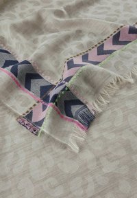 Hellbeige Schal mit subtilen Tiermotiven und einem lebhaften Chevron-Muster-Rand in marineblau, rosa und grünen Akzenten sowie fransigen Kanten.