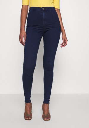 Kobieta ubrana w ciemnoniebieskie, obcisłe dżinsy z wysokim stanem i oliwkowo-zielone sandały na obcasie z paskami, połączona z żółtym prążkowanym topem, stoi na białym tle.