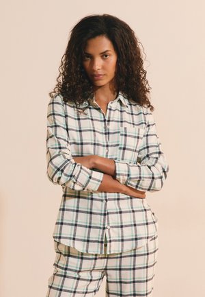 Femme aux cheveux bouclés portant une chemise à carreaux verte et blanche boutonnée et un pantalon assorti, debout les bras croisés devant un fond beige.