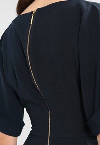 Granatowa sukienka z dekoltem łódka i krótko rolowanymi rękawami, z widocznym złotym zamkiem biegnącym pionowo wzdłuż pleców.