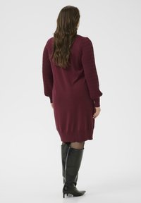 Weinrotes Strickkleid mit gerippten Ärmeln und einem Saum knapp über dem Knie, kombiniert mit schwarzen kniehohen Stiefeln. Glatte Textur im gesamten Kleid.