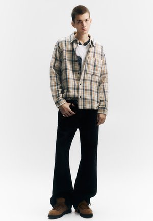 Cămașă din flanel în carouri beige, albastru și maro, asortată cu blugi negri cu picioare largi și pantofi maro cu șireturi și finisaj texturat.