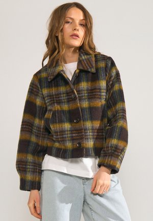 Junge Frau trägt eine braun-gelb-grün karierte, geknöpfte Jacke über einem weißen Hemd und hellblauen Jeans, steht vor einem schlichten Hintergrund.