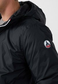 Giacca nera imbottita con cappuccio, caratterizzata da una texture liscia, un cordoncino elastico e un patch circolare logo sulla manica.