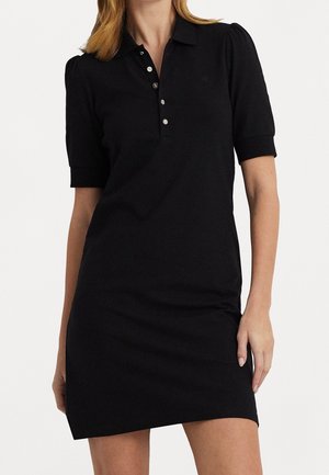 Femme portant une robe polo noire ajustée à manches courtes avec un col boutonné et des poignets côtelés, debout devant un fond uni.