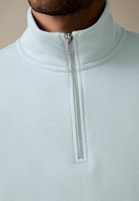 Lichtblauwe pullover met een hoge kraag en halve rits. Zacht, gestructureerd materiaal met contrasterende zilveren hardware.