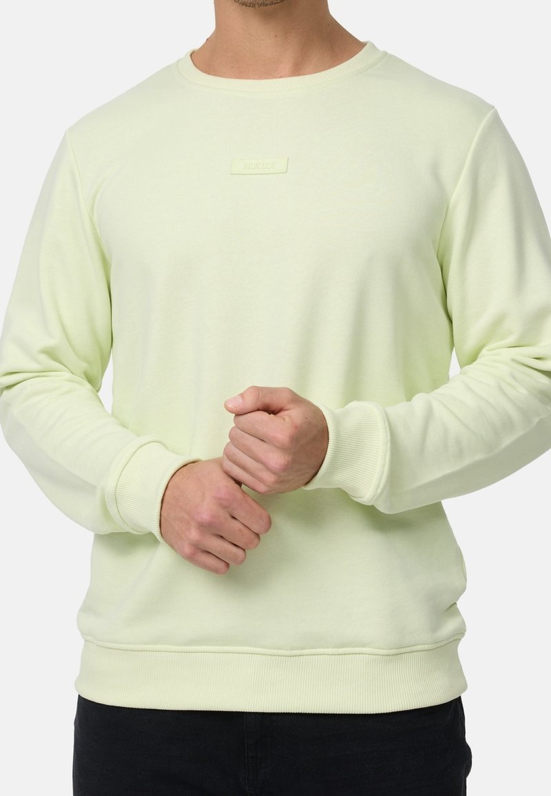 Homme portant un sweat-shirt jaune pâle à manches longues avec poignets et ourlet côtelés, mains jointes devant, torse visible, fond uni.