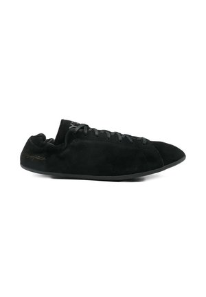 Sneaker minimalista in camoscio nero con lacci neri, suola piatta e collo elastico alla caviglia, mostrata di profilo su sfondo bianco.
