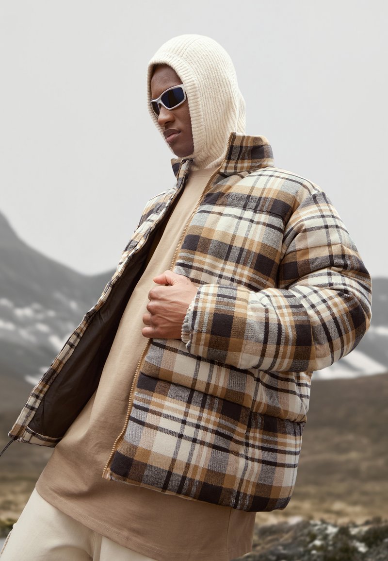 Padded jacket w kratkę w odcieniach brązu, beżu i bieli; ma wysoki kołnierz; noszony na beżowej koszuli; model w dzianinowym kapturze i okularach przeciwsłonecznych.