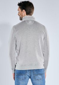 Helles graues Sweatshirt mit hohem Kragen und gerippten Bündchen, mit einem Etikett auf der Rückseite. Kombiniert mit Jeans aus Denim, die Details der Gesäßtaschen zeigen.