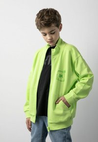 Neon zielona bluza z kapturem na zamek błyskawiczny, z wysokim kołnierzem, akcentem logo na klatce piersiowej i bocznymi kieszeniami. Noszona na czarnej koszulce i niebieskich dżinsach.