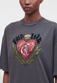 Κοντινή λήψη ενός σκούρου γκρι T-shirt με κόκκινη καρδιά, πράσινα φύλλα, κορώνα και το κείμενο "Tired Earth" καθώς και διάφορα μικρά γράμματα.