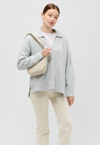 Maglione grigio chiaro lavorato a maglia con scollo a V e polsini a costine, abbinato a pantaloni color crema. La modella tiene una borsa a tracolla beige.