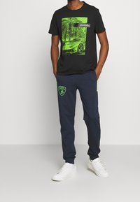 Černé tričko s neonově zelenou grafikou Lamborghini a palmami. Námořnické modré joggers s zeleným logem. Bílé tenisky dokončují outfit.