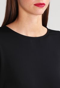 Nainen, jolla on pitkät ruskeat hiukset, pukeutuneena mustaan pyöreäkaulaiseen puseroon ja kirkkaan punaiseen huulipunaan, kuvattuna hartioista huuliin vaaleaa taustaa vasten.