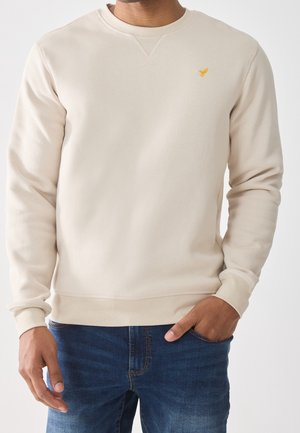 Man wearing a beige long-sleeve sweatshirt with a small yellow bird logo on the chest and blue jeans, one hand in pocket.