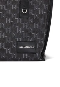 Svart texturerad väska med ett upprepande grått logomönster. Har en läderetikett med "KARL LAGERFELD" präglat i silver. Metallbeslag som accent.