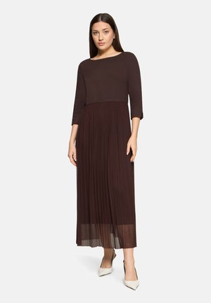 Frau steht auf einfachem Hintergrund, trägt ein dunkelbraunes langärmliges Oberteil und einen passenden plissierten Maxirock sowie weiße spitze Absätze.