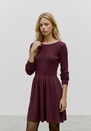 Donna bionda con un vestito di maglia bordeaux a maniche lunghe, in piedi davanti a uno sfondo grigio chiaro uniforme.