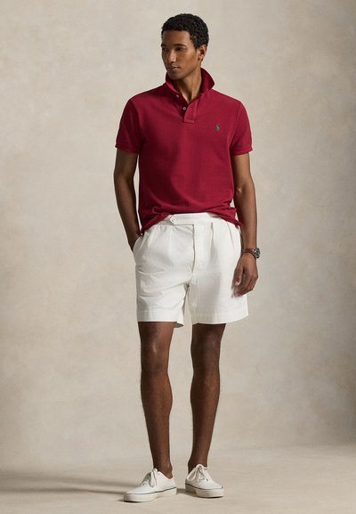 Man wearing red polo shirt, white pleated shorts, white sneakers, and a wristwatch, standing with hands in pockets against plain background.