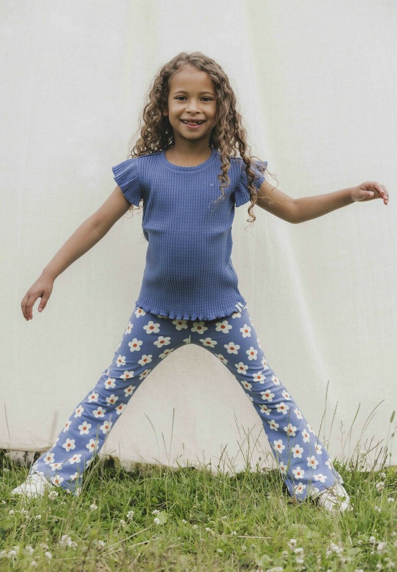 Blauwe geribde top met korte mouwen en een rucheszoom, gecombineerd met lichtblauwe flared broek met een wit bloemenpatroon. Witte sneakers.