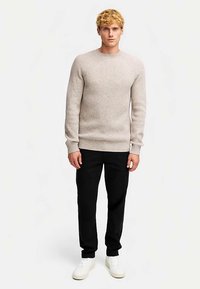 Lys beige ribbestrikket genser med lange ermer, rund hals, kombinert med svarte bukser og hvite joggesko. Enkelt, strukturert design.