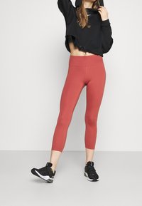 Leggings rouges coupés en tissu extensible, associés à un crop hoodie noir. Présentent une taille haute et une texture lisse. Chaussures de sport noires.