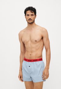 Boxers bleu clair avec une ceinture élastique rouge portant le logo "HUGO", fermeture à un bouton, et détails de couture subtils.