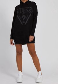 Žena v černém oversized mikinovém šatech s velkým logem "Guess" v kombinaci s bílými chunky teniskami, stojící na jednotném pozadí.