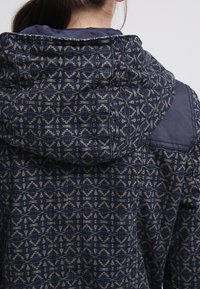 Marinblå jacka med geometrisk mönster i ljusbeige, med huva och textilmaterial. Inkluderar marinblå axeldetaljer.