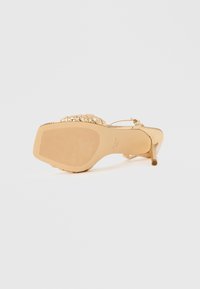 Χρυσό πέδιλο με υφασμένο πάτο, διακοσμημένα μεταλλικά στοιχεία στο πάνω μέρος και λεπτό λουράκι στον αστράγαλο για στήριξη.