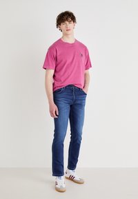 Roze katoen T-shirt met korte mouwen, gecombineerd met donkerblauwe slim-fit jeans. Witte schoenen met rode strepen en bruine zolen. Eenvoudige achtergrond.