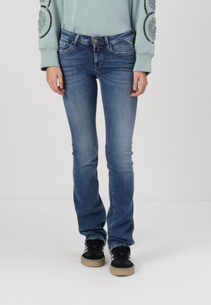 Osoba ubrana w wyblakłe niebieskie dzwonowe dżinsy, czarne trampki oraz jasnoniebieską bluzę z haftowanymi detalami na rękawach.