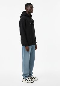 Felpa nera con cappuccio e tasca frontale, con testo bianco. Indossata con jeans dritti azzurri e scarpe da ginnastica nere e bianche.